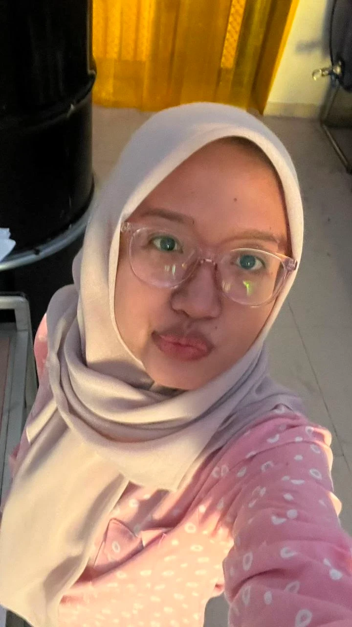 Caca in a light hijab and pink shirt, taking a selfie in warm light
