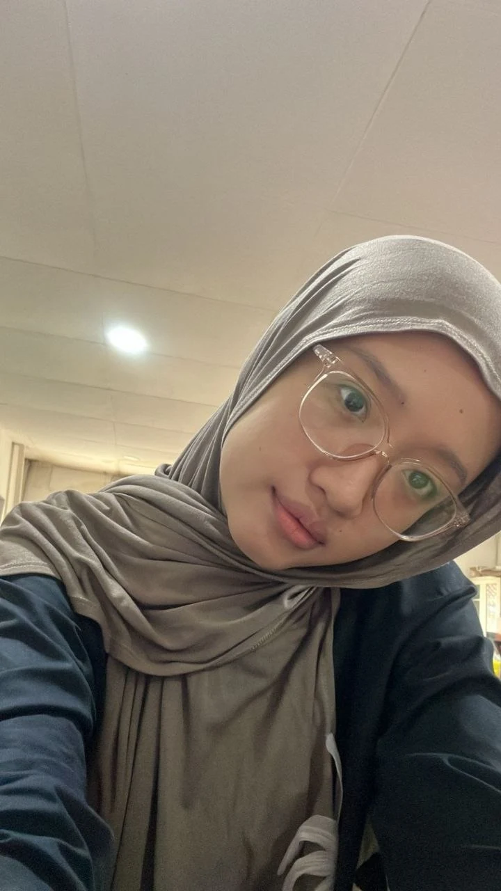 Caca wearing glasses and a taupe hijab, tilting her head for a selfie